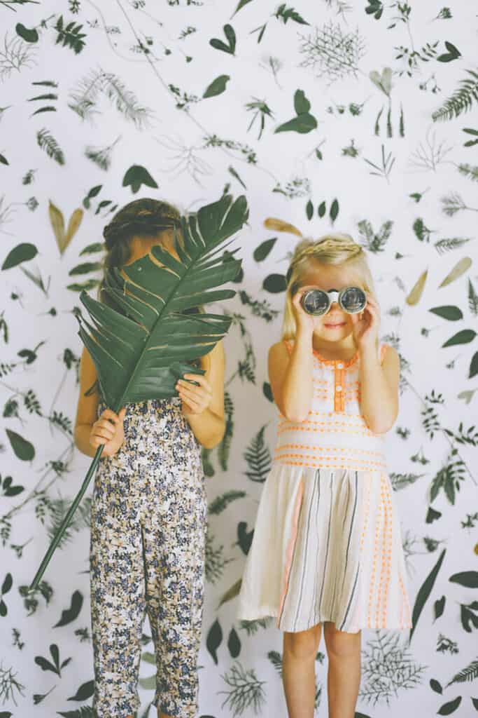 two young girls standing in front of a wall with tropical wall paper holding tropical leaves