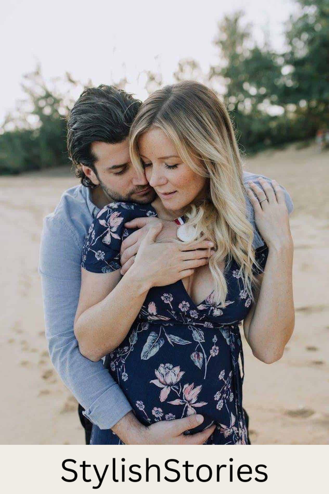 image of a man standing behind a pregnant woman on the beach giving her a hug, she is pregnant and wearing a blue floral dress