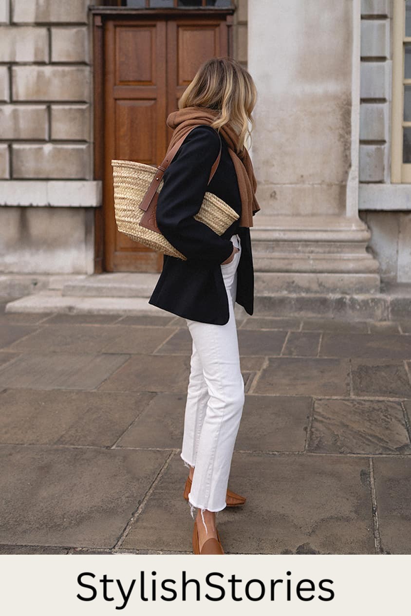 Woman wearing white jeans, tan loafers, a black blazer and a brown scarf walks through the city.