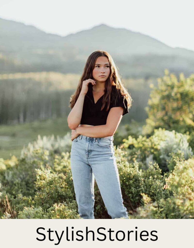 wholesome image of a teen girl with brown hair wearing a black t-shirt and jeans standing in a field with mountains in the background for senior photos