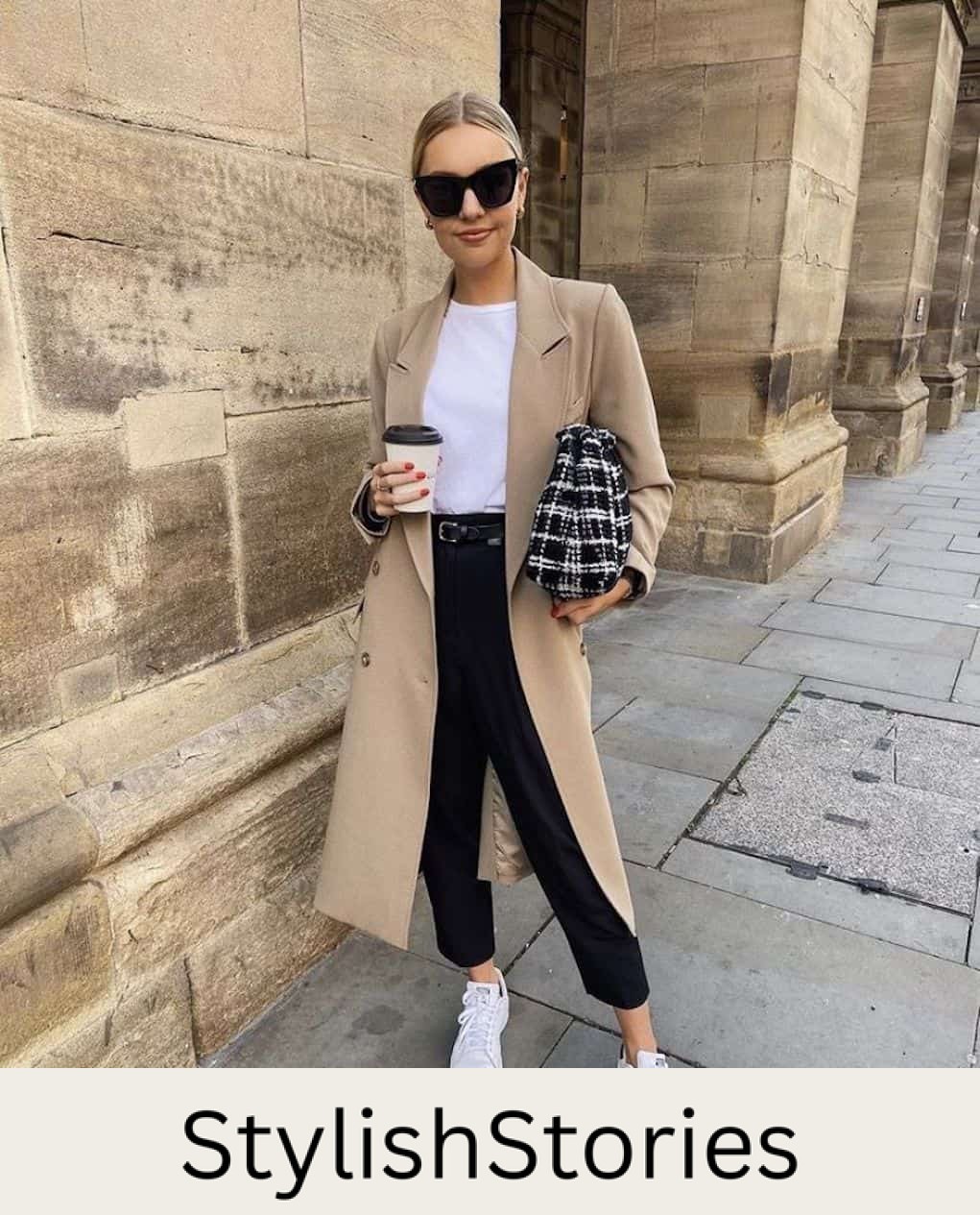 Woman wearing a tan trench coat, black jeans, white sneakers and a white t-shirt sips coffee in the city.