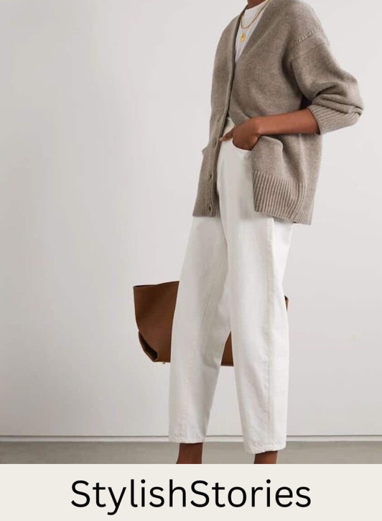 Woman wearing white jeans, black sandals, and grey sweater poses in front of a white wall.
