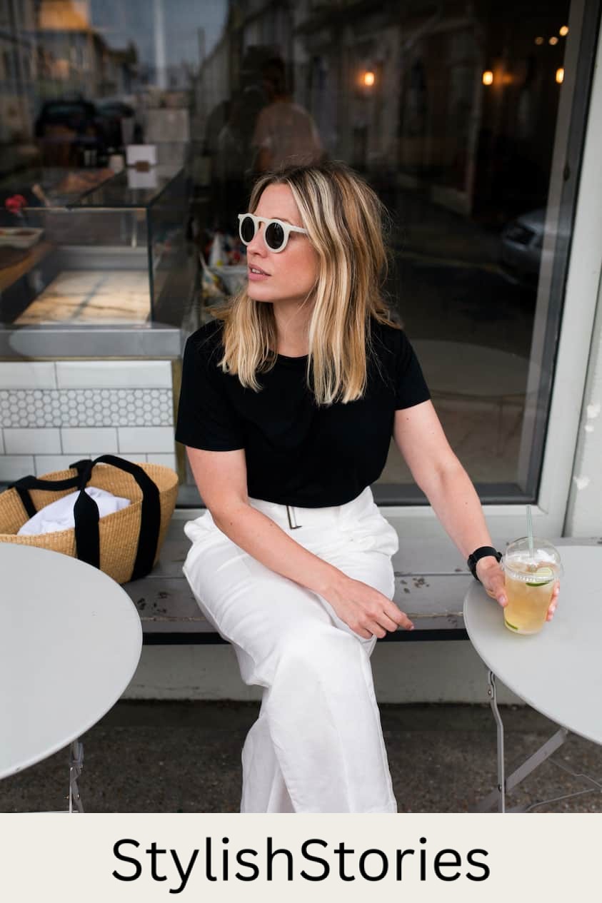 Woman wearing white linen pants, a black t-shirt and white sunglasses sips on a drink while sitting on a patio.