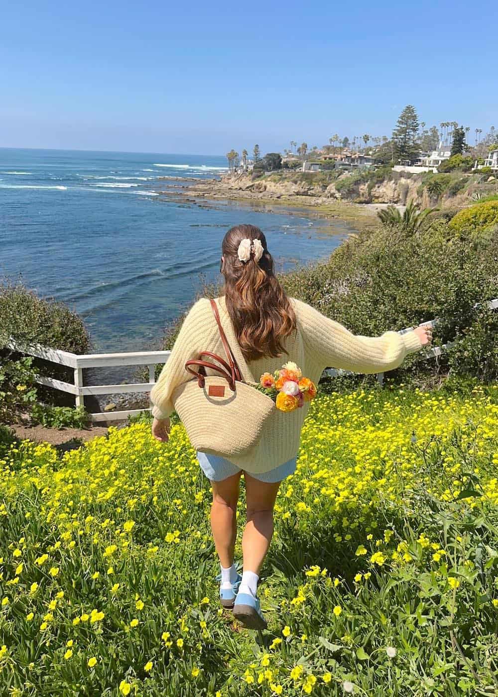a woman wearing a butter yellow cardigan and light blue shorts with a raffia tote and blue sneakers