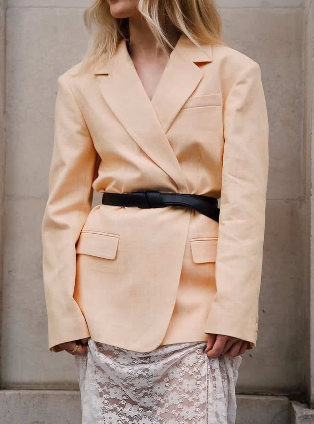 Woman wearing a beige blazer and lace skirt with a chunky black belt.
