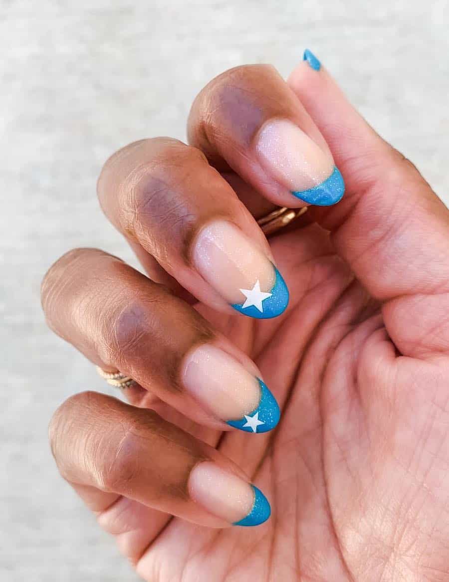 short nude almond nails with blue tips, a glitter topcoat, and white star accents