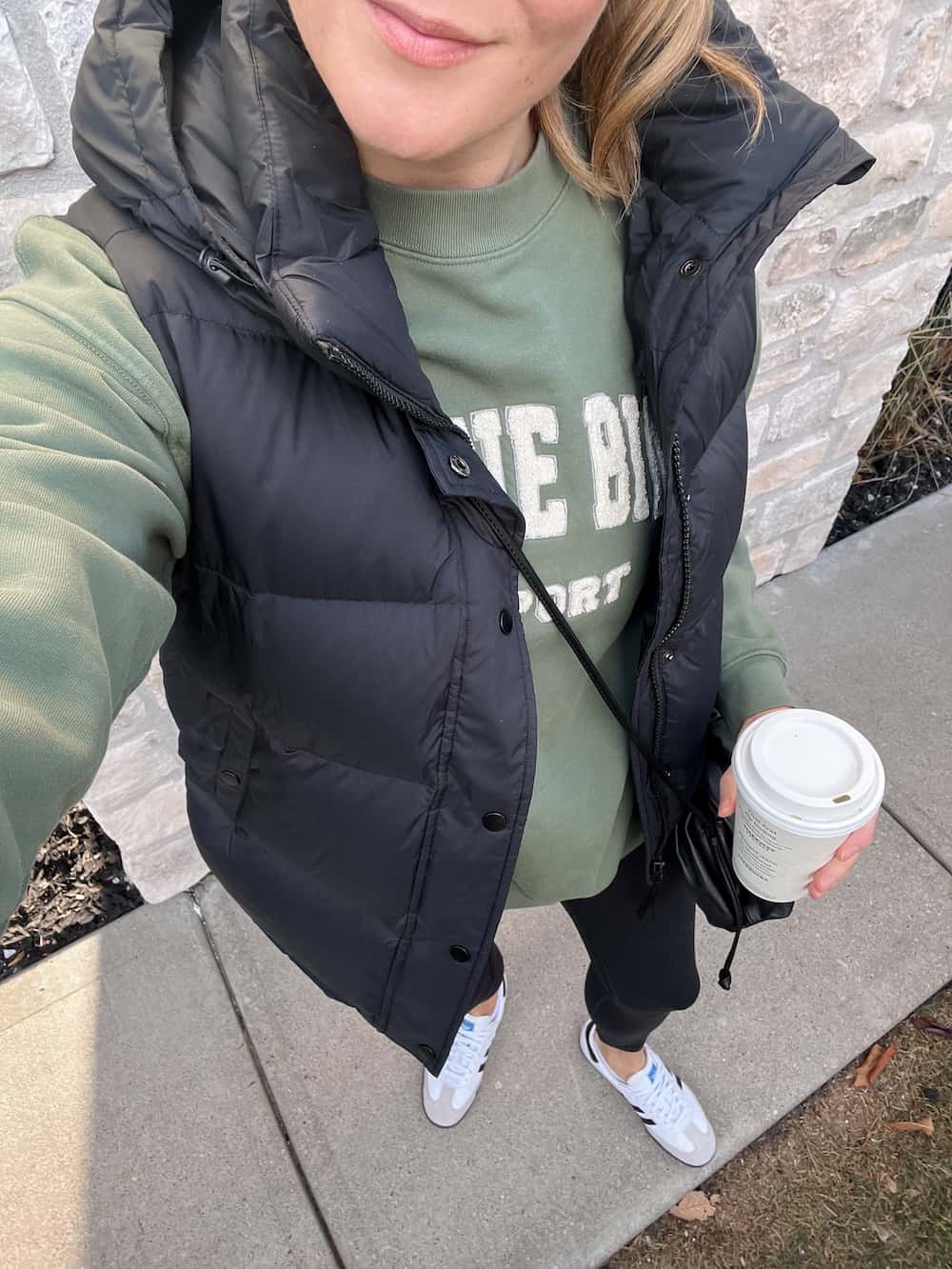 woman wearing a black puffer vest that is a dupe of the Aritzia Super Puff vest over a green sweatshirt with green leggings and white sneakers