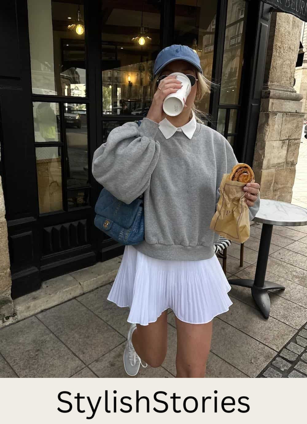 Woman wearing a white pleated tennis skirt with a grey crew neck sweatshirt and a blue baseball cap.
