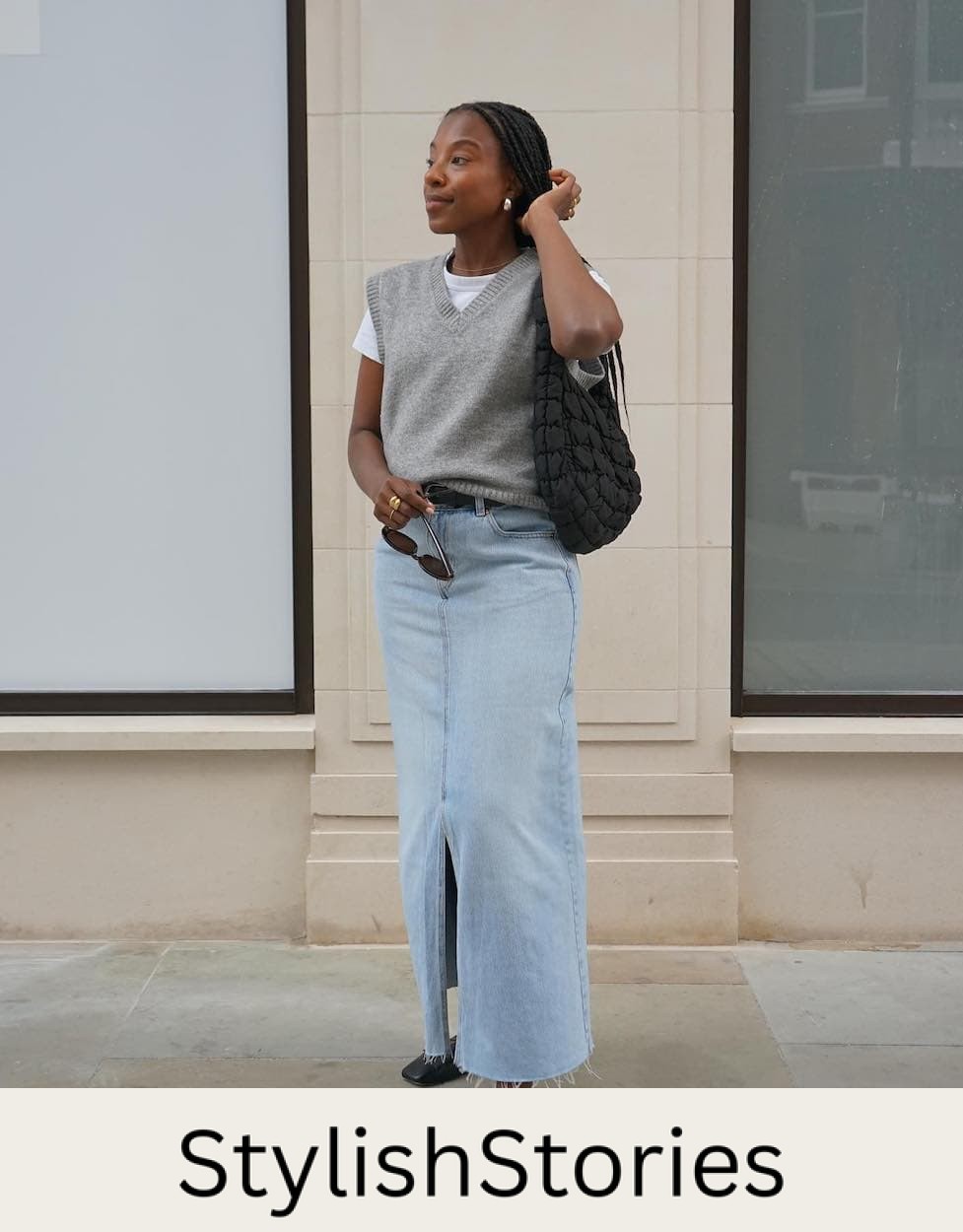 Woman wearing a denim midi skirt with a whit t-shirt, a grey sweater vest and black ballet flats.