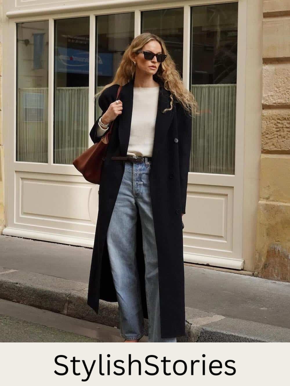 Woman wearing baggy jeans, pointed toe pumps, a cream sweater and an oversized black trench coat.