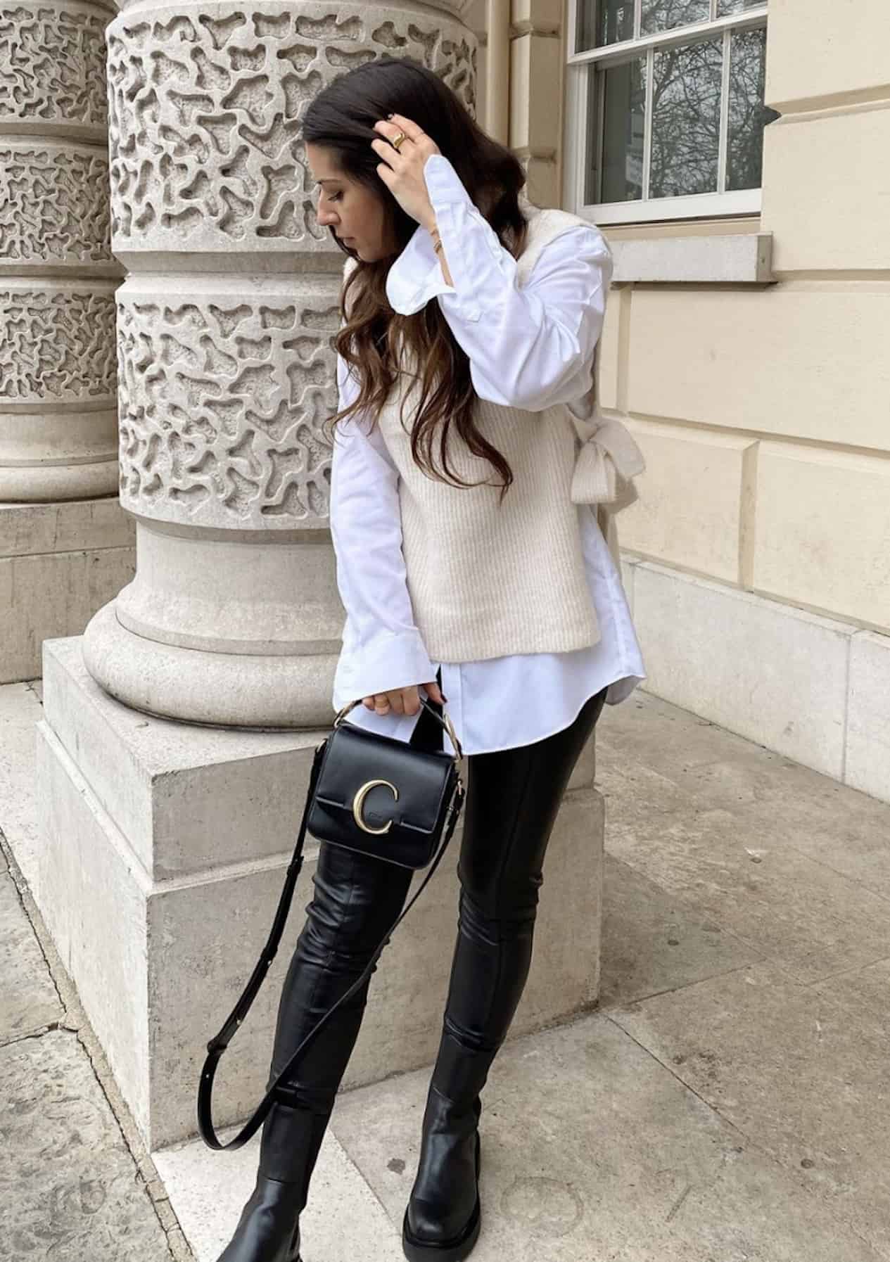 image of a woman wearing a white collared shirt, cream knit vest, black leather leggings, and black boots