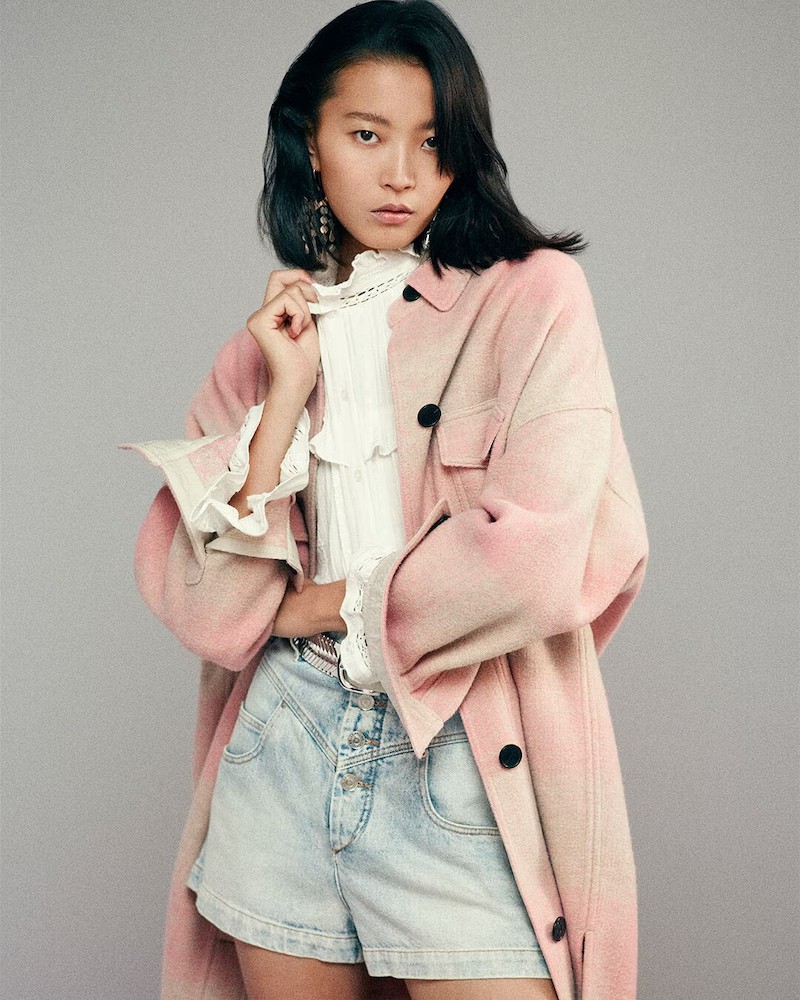 Woman wearing a light wash denim shorts, a white blouse and a pink and cream shacket.