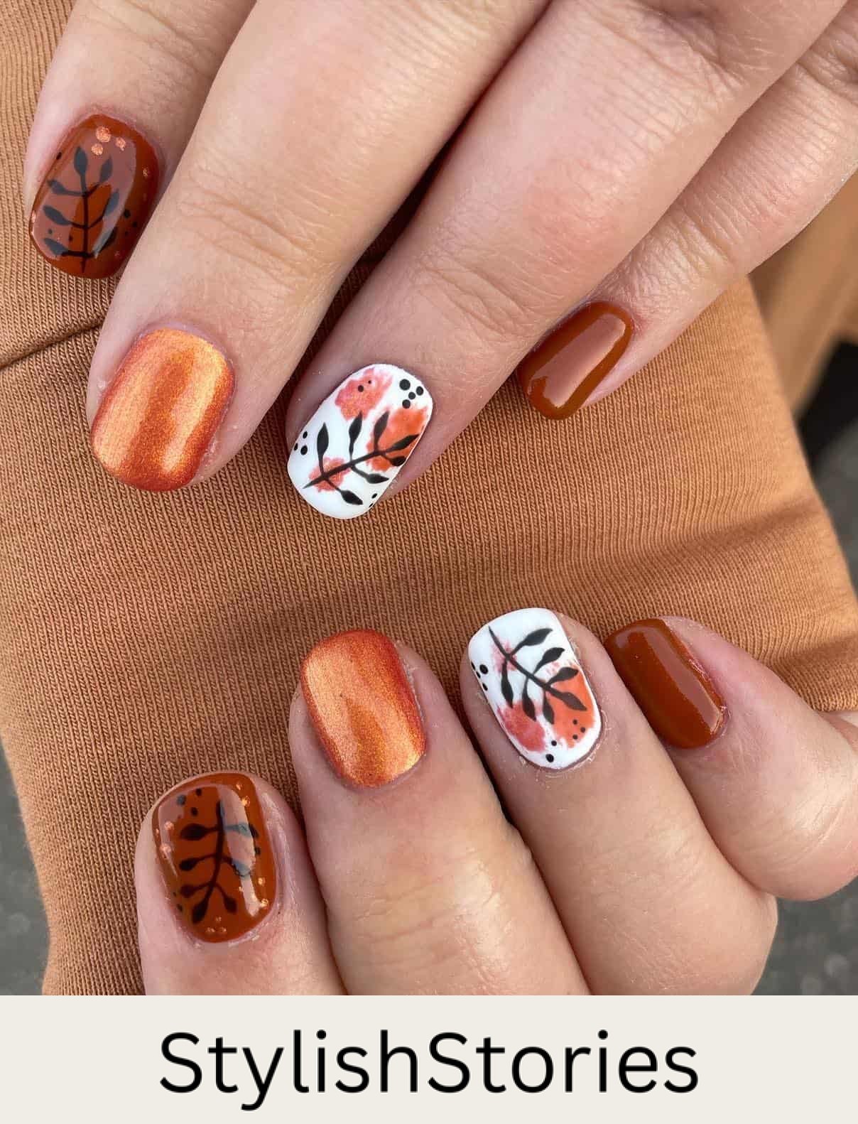 a hand with short nails painted in shimmering burnt orange, glossy brown, and glossy white polish with leaf nail art