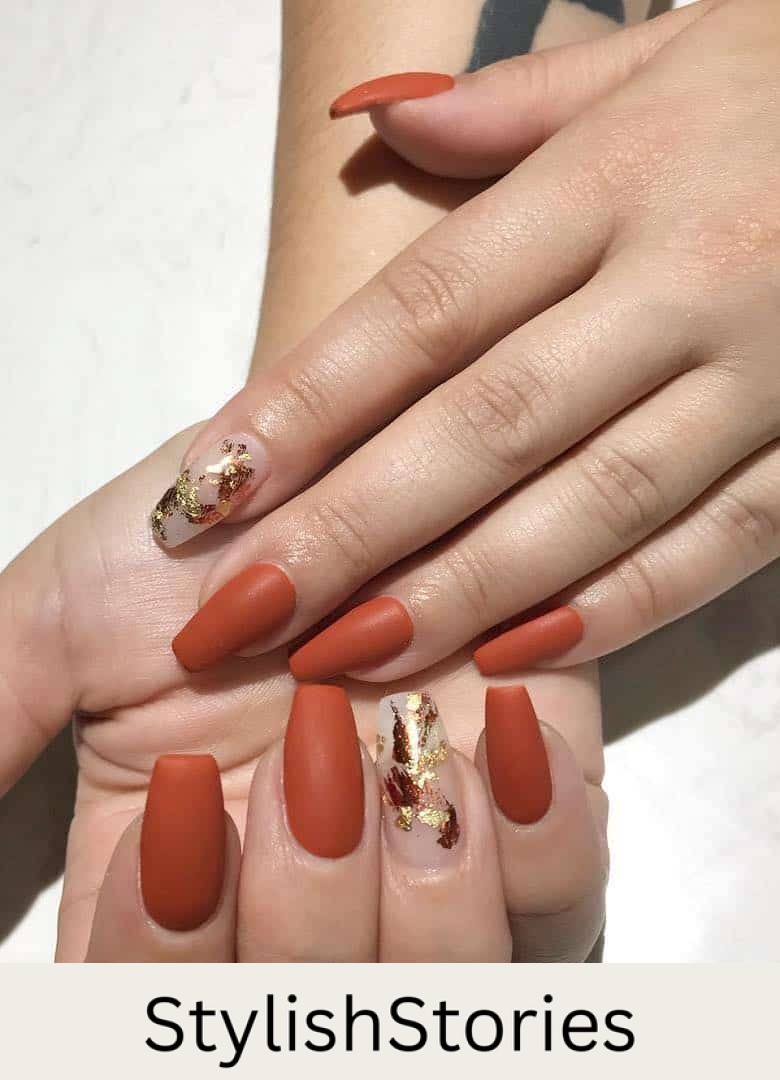 a hand with medium-length coffin nails painted a matte burnt orange shade with nude accent nails with gold foil