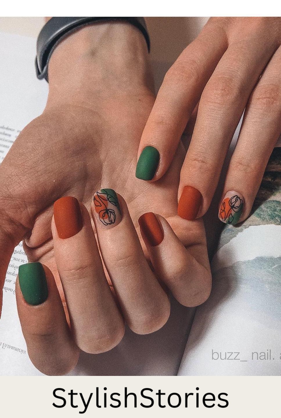 a hand with short square nails painted in matte dark green and burnt orange with a nude accent nail featuring both shades and black floral line art