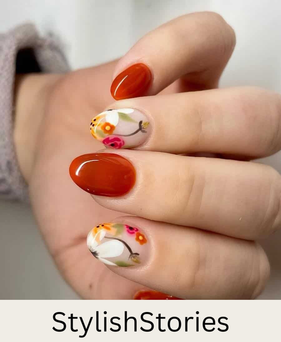 a hand with short round nails painted solid-colored burnt orange and nude glossy accent nails with colorful floral nail art