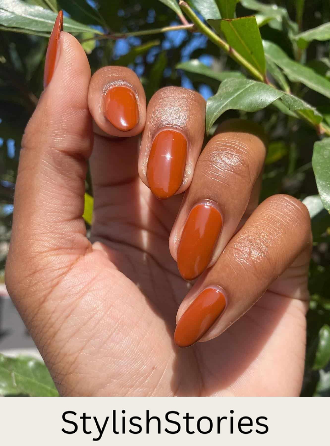 a hand with medium round nails painted with a burnt orange polish and gloss finish