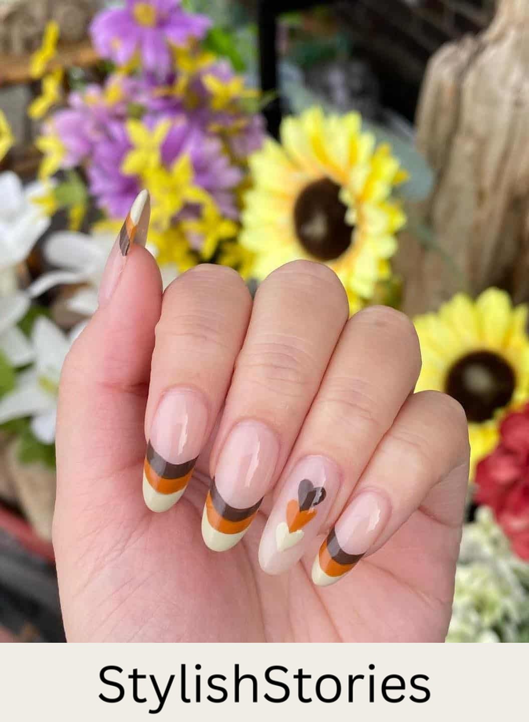 a hand with long round nails painted in cream, burnt orange, and brown French tips with a heart accent nail