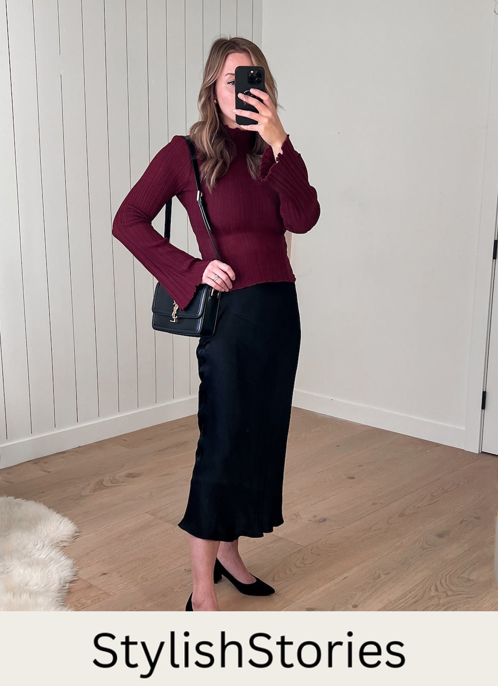 A woman wearing a black silk midi skirt with a red knit crewneck sweater, black heels, and a black shoulder bag