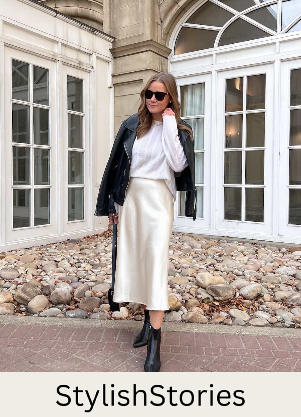 A woman wearing an ecru colored silk midi skirt with a white knit sweater, a black leather jacket, and black leather ankle boots