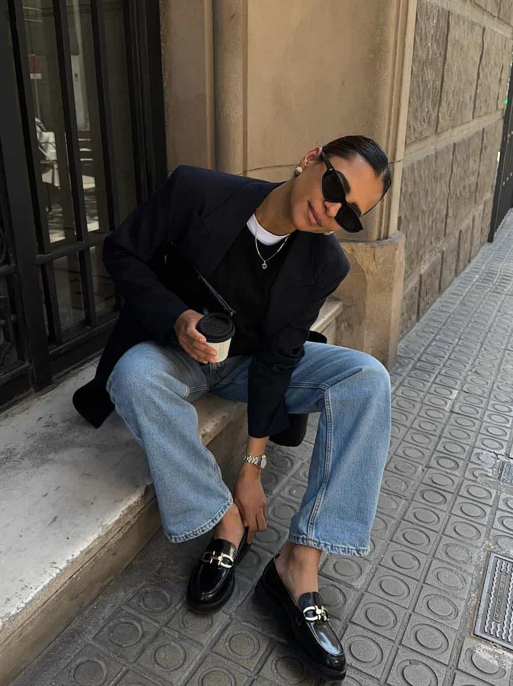 a woman wearing chunky black loafers, straight-leg blue jeans, a black tee, and a black blazer