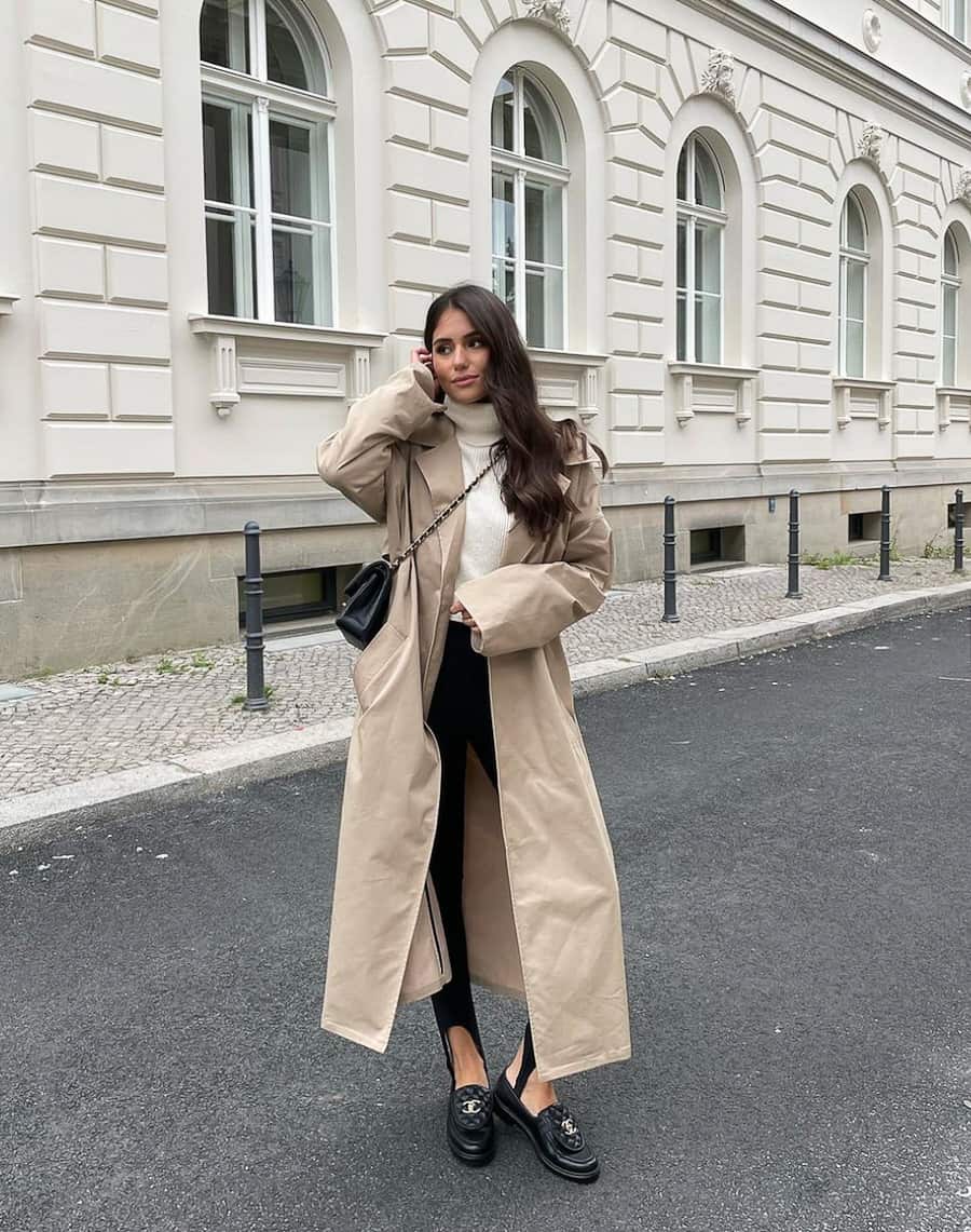 image of a woman wearing a long trench coat over a knit turtleneck sweater, black stirrup leggings, and black loafers