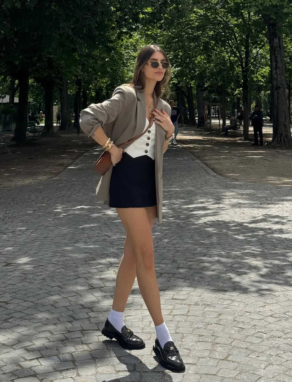 a woman wearing a black mini skirt, a white vest, a medium brown blazer, and chunky black loafers
