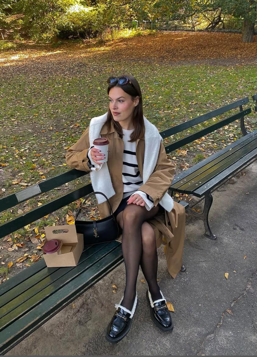 Woman wearing a black skirt with tights, a white and black stripped sweater, a tan trench coat and black loafers.