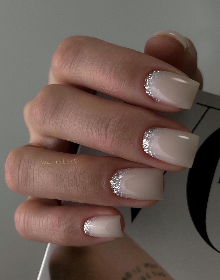 A hand with short square nails painted a milky cream color with silver glitter reverse French tips