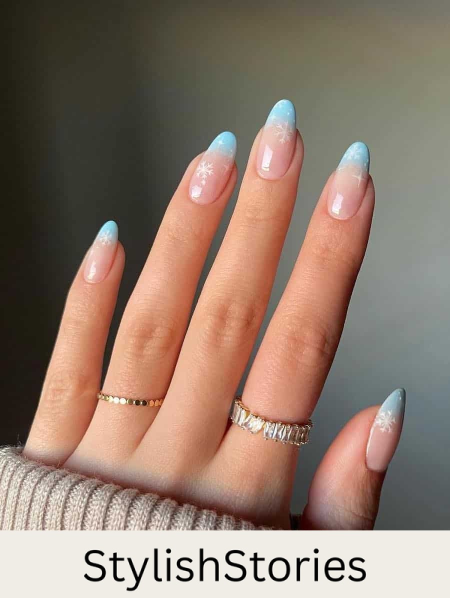 A hand with medium almond nails painted a light blue and nude ombre with white snowflakes