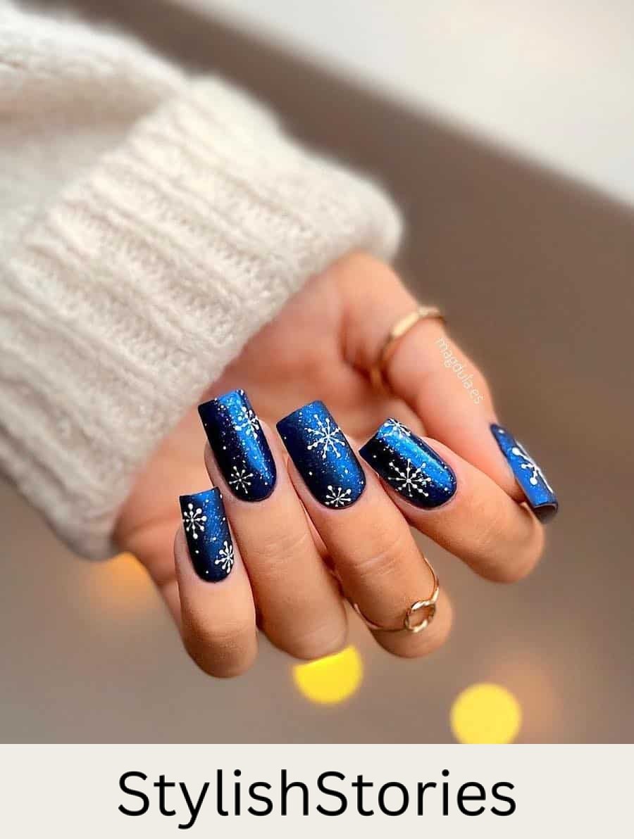 A hand with long square nails painted a shimmering dark blue with white splatter accents and snowflakes