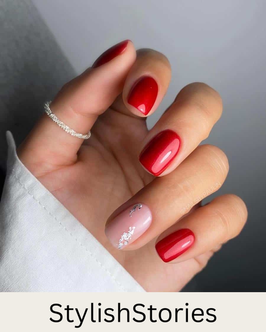 A hand with short squoval nails painted in red polish with one nude accent nail with silver flakes