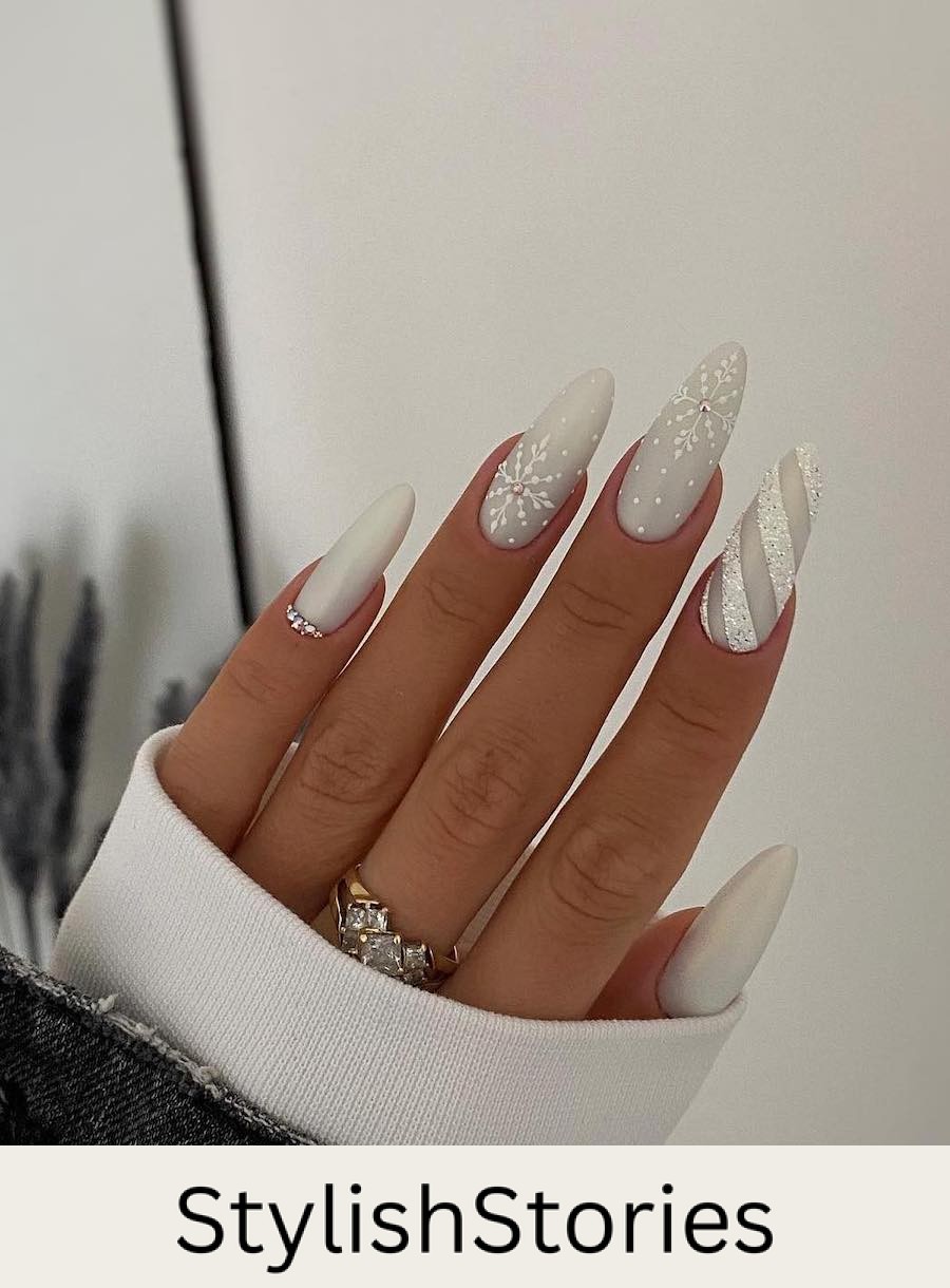 A hand with long white almond nails painted with multiple designs, including shimmering white stripes, snowflake art, and crystal accents