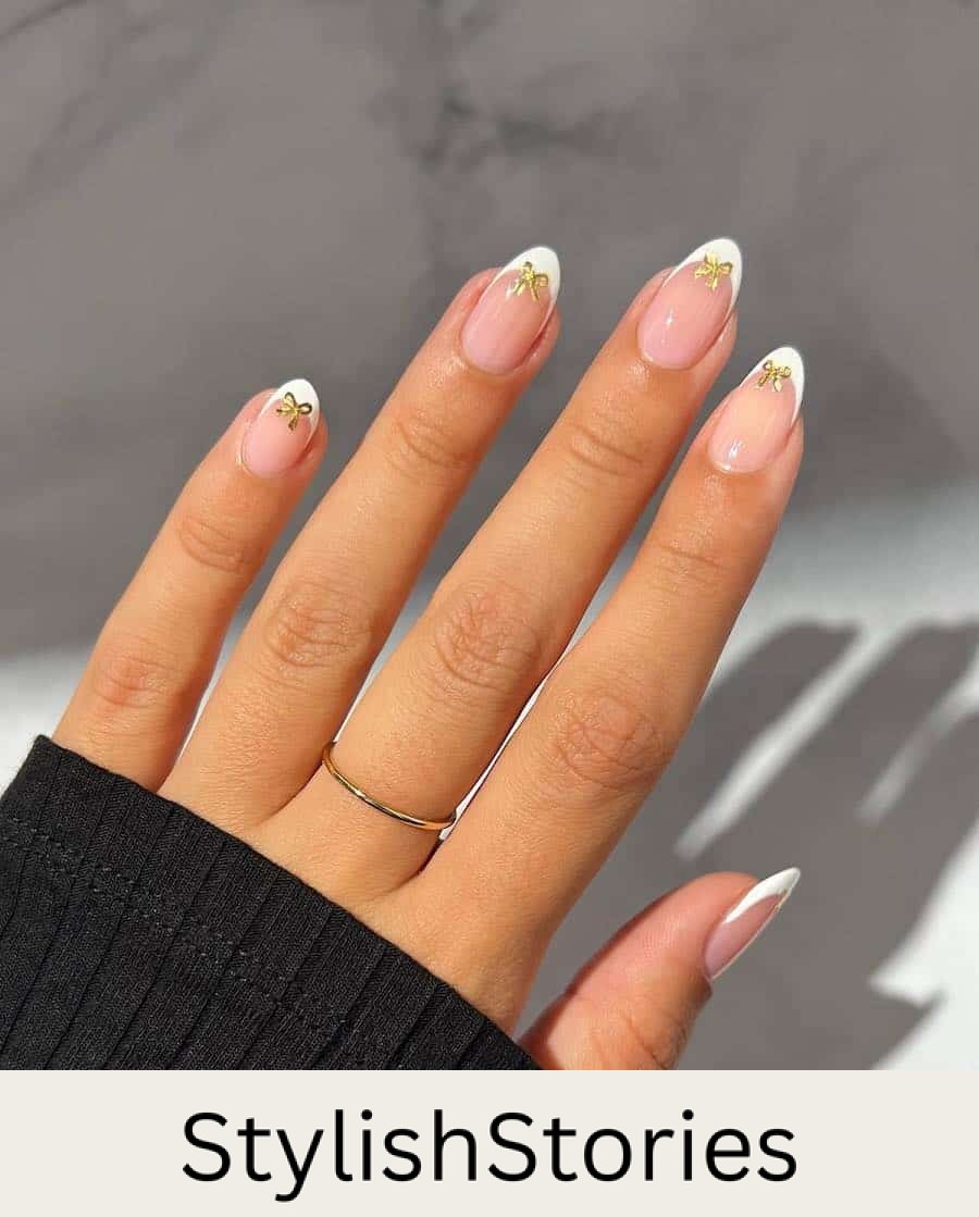 A hand with short almond nails painted with white French tips on nude pink nails with gold bow details