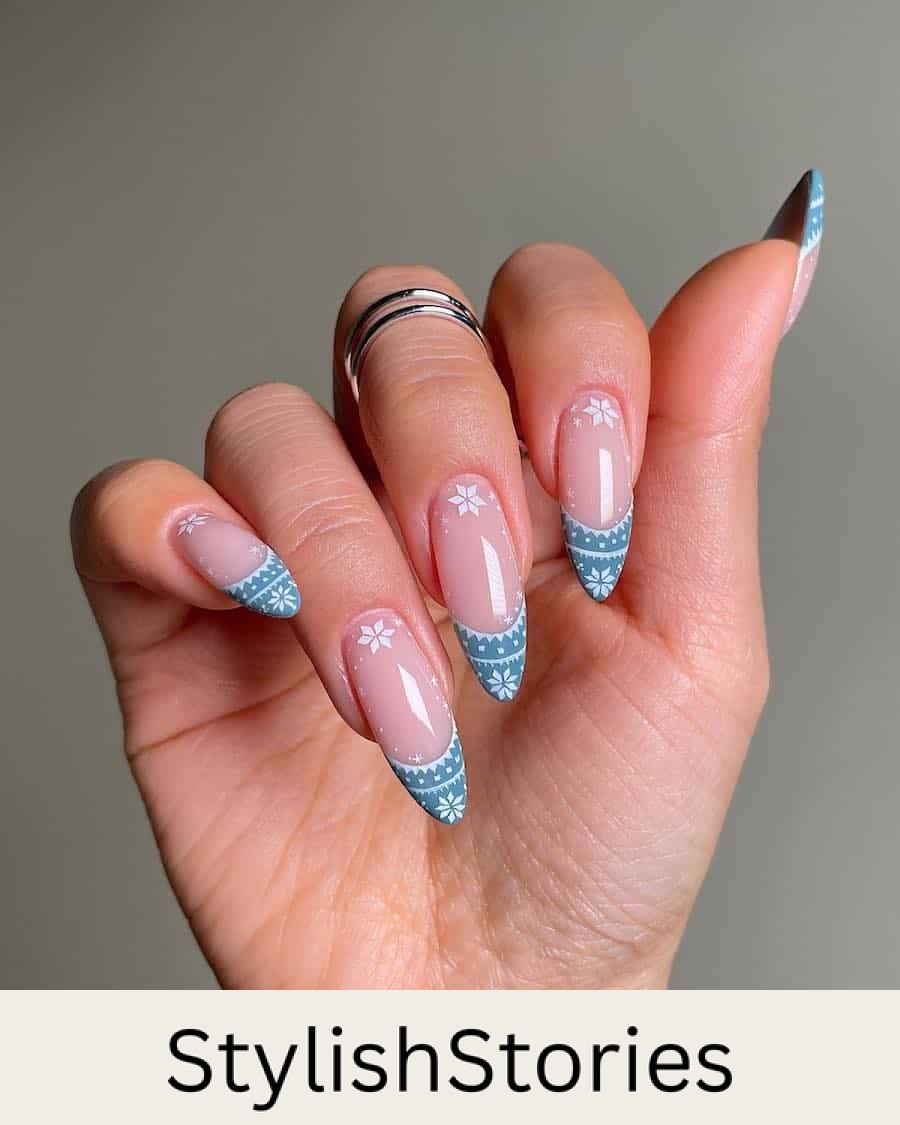 A hand with long almond nails painted a glossy nude pink with blue and white sweater detailed French tips and white snowflake accents