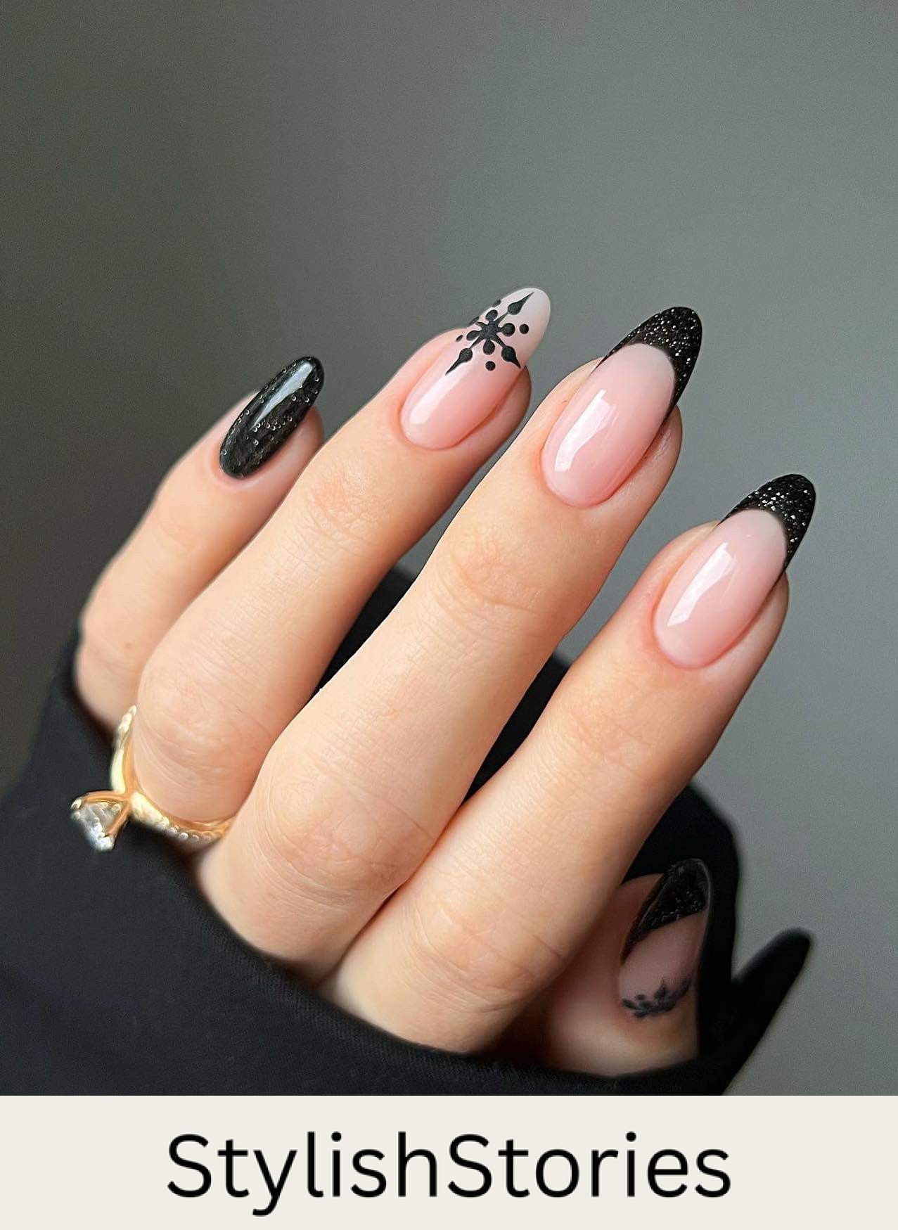 A hand with medium nude almond nails with black glitter French tips, a black glitter accent nail, and black snowflake details