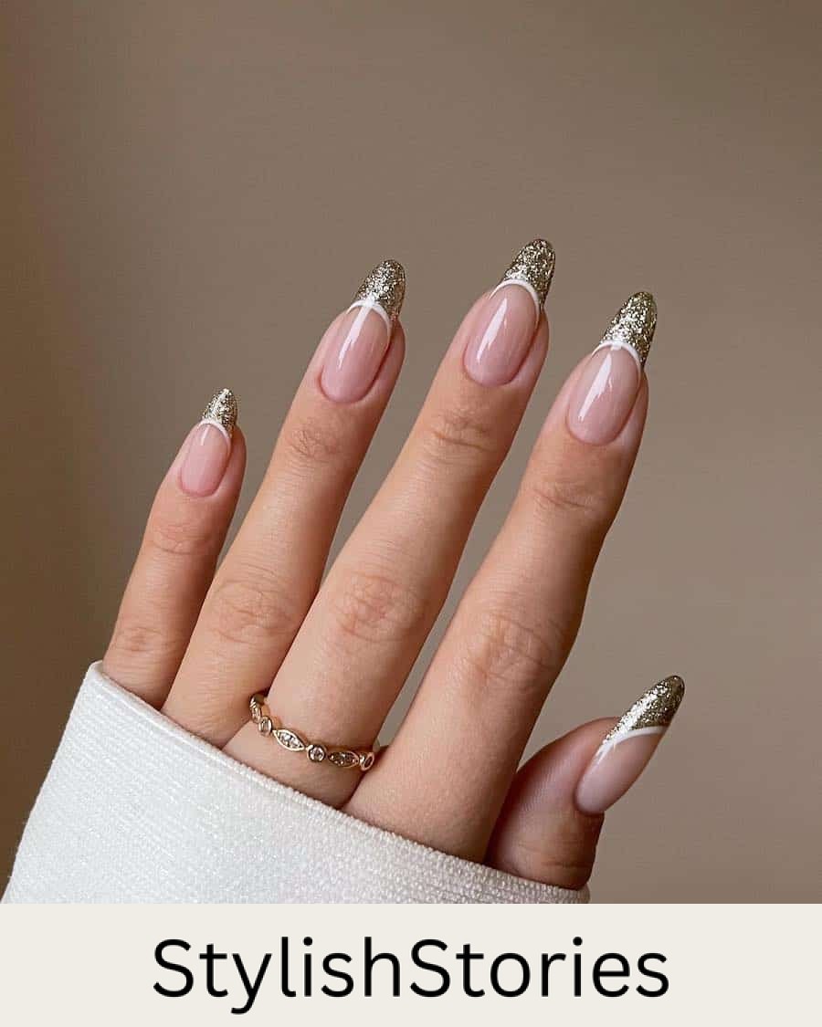 A hand with long glossy nude almond nails with gold glitter French tips and white borders