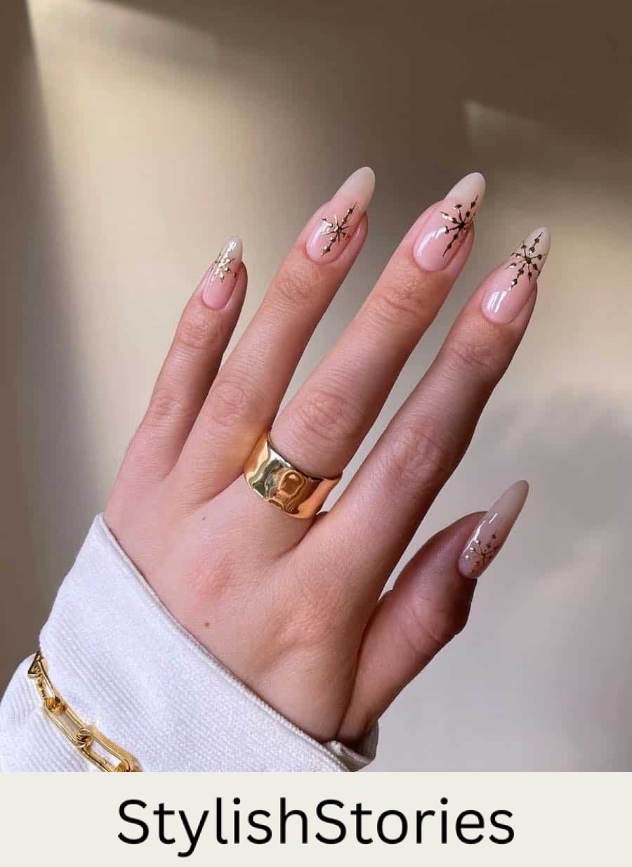 A hand with long nude almond nails with gold snowflakes