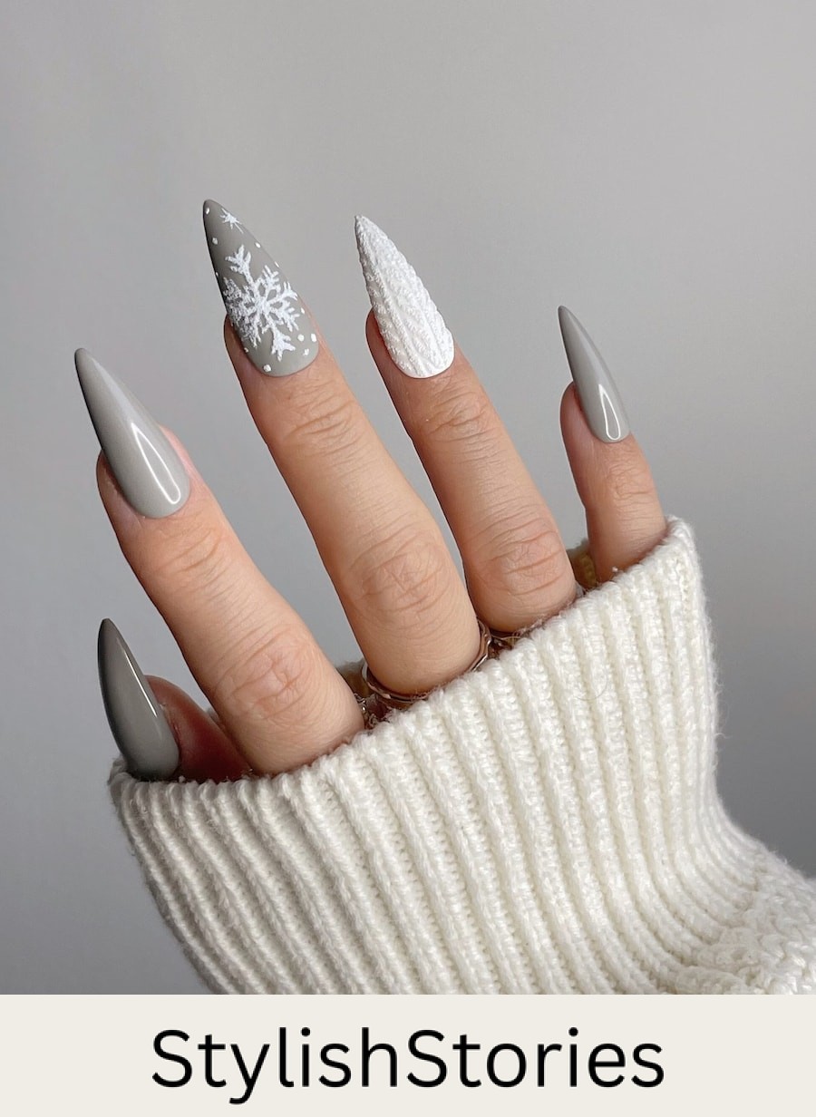 A hand with long stiletto nails painted a glossy grey color with a white sweater accent nail and a matte grey nail with white snowflakes