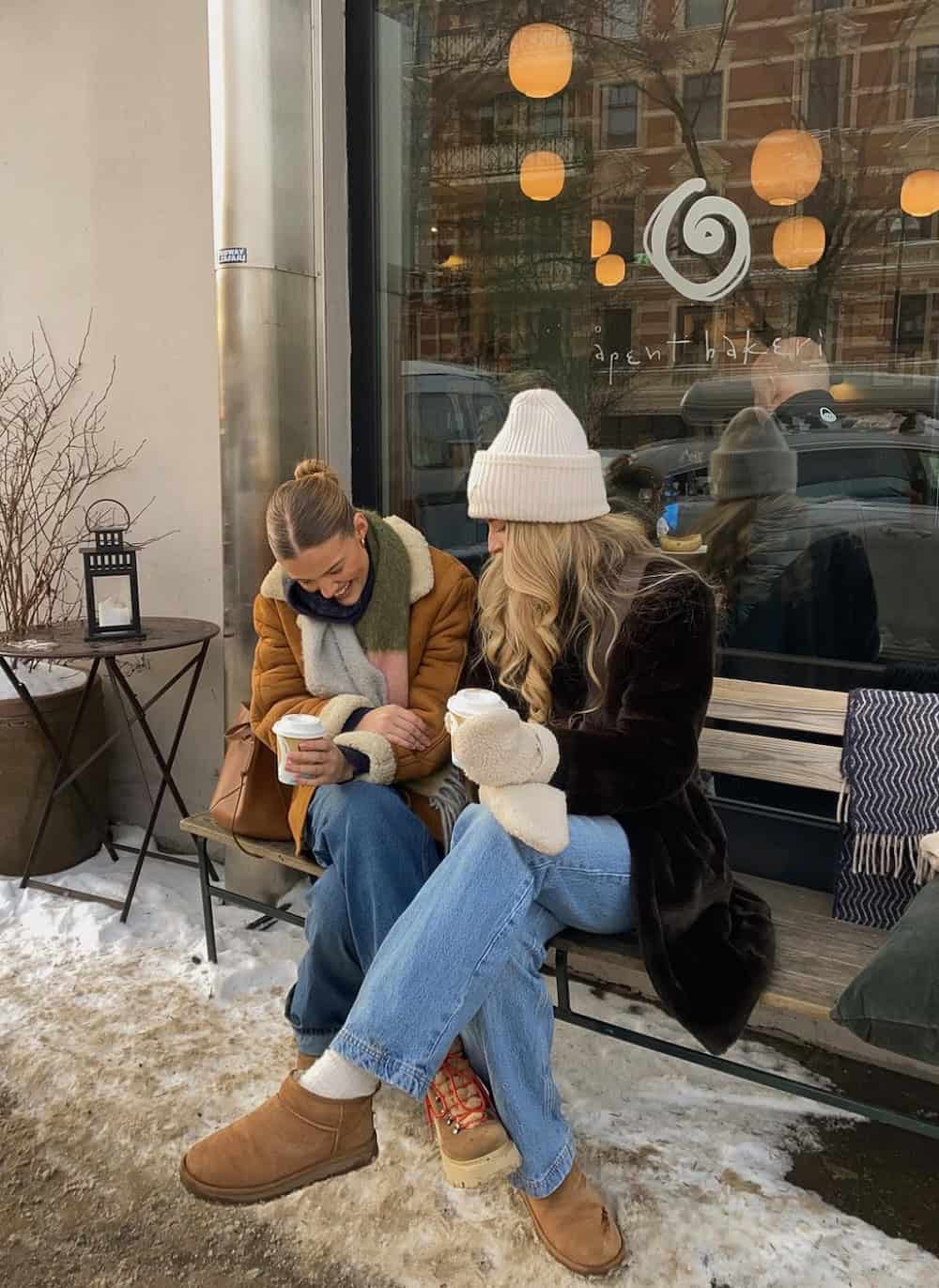 Two women wearing jeans and coats sipping coffee outside a cafe.