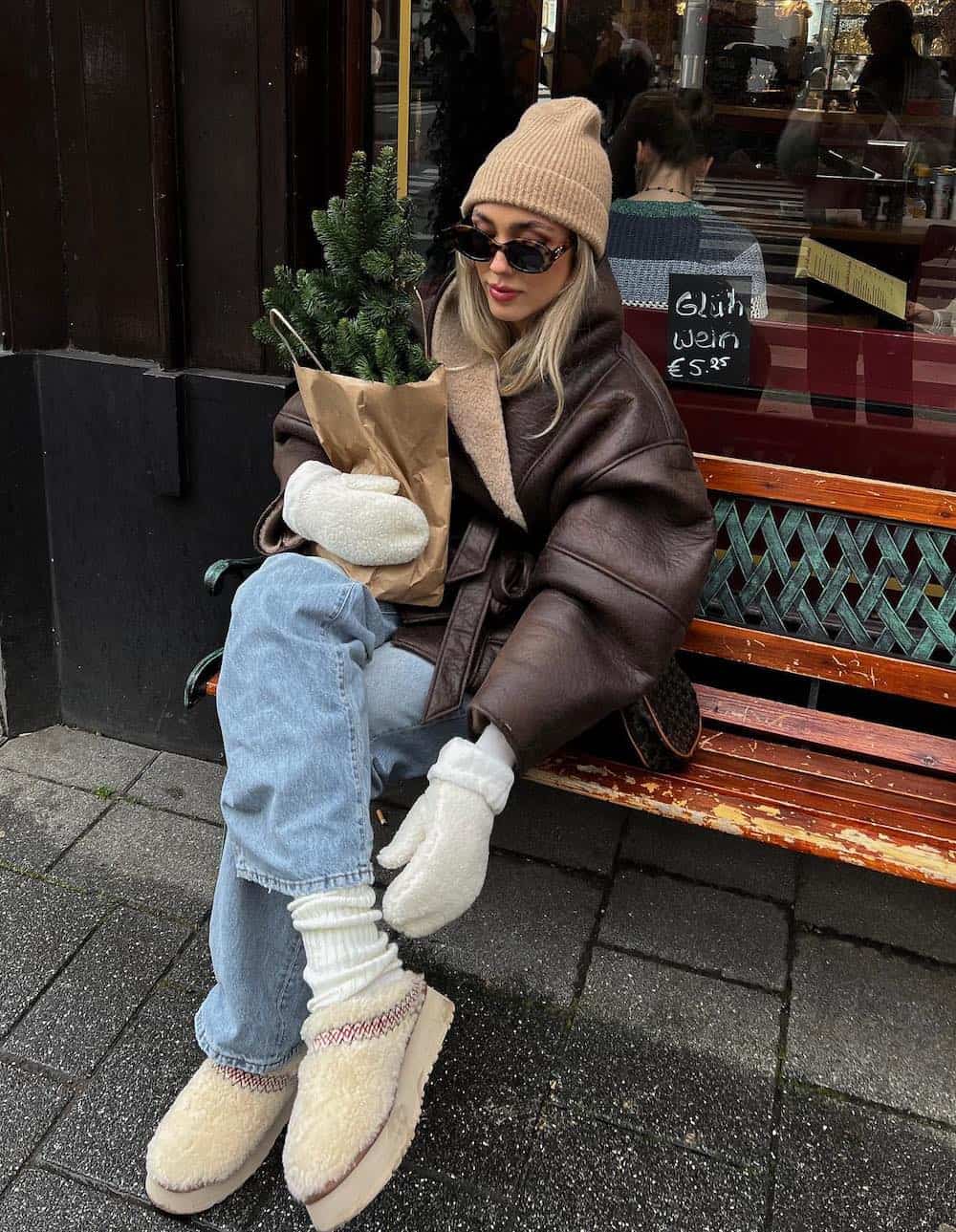 Woman wearing jeans with a brown leather jacket, a tan beanie and sherpa Ugg slippers.