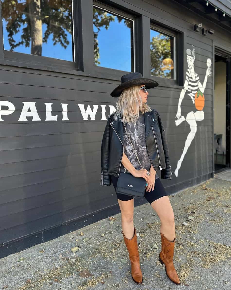 An image of a woman with an edgy country concert outfit featuring running shorts, a graphic tee, a leather jacket, and a cowboy hat and boots