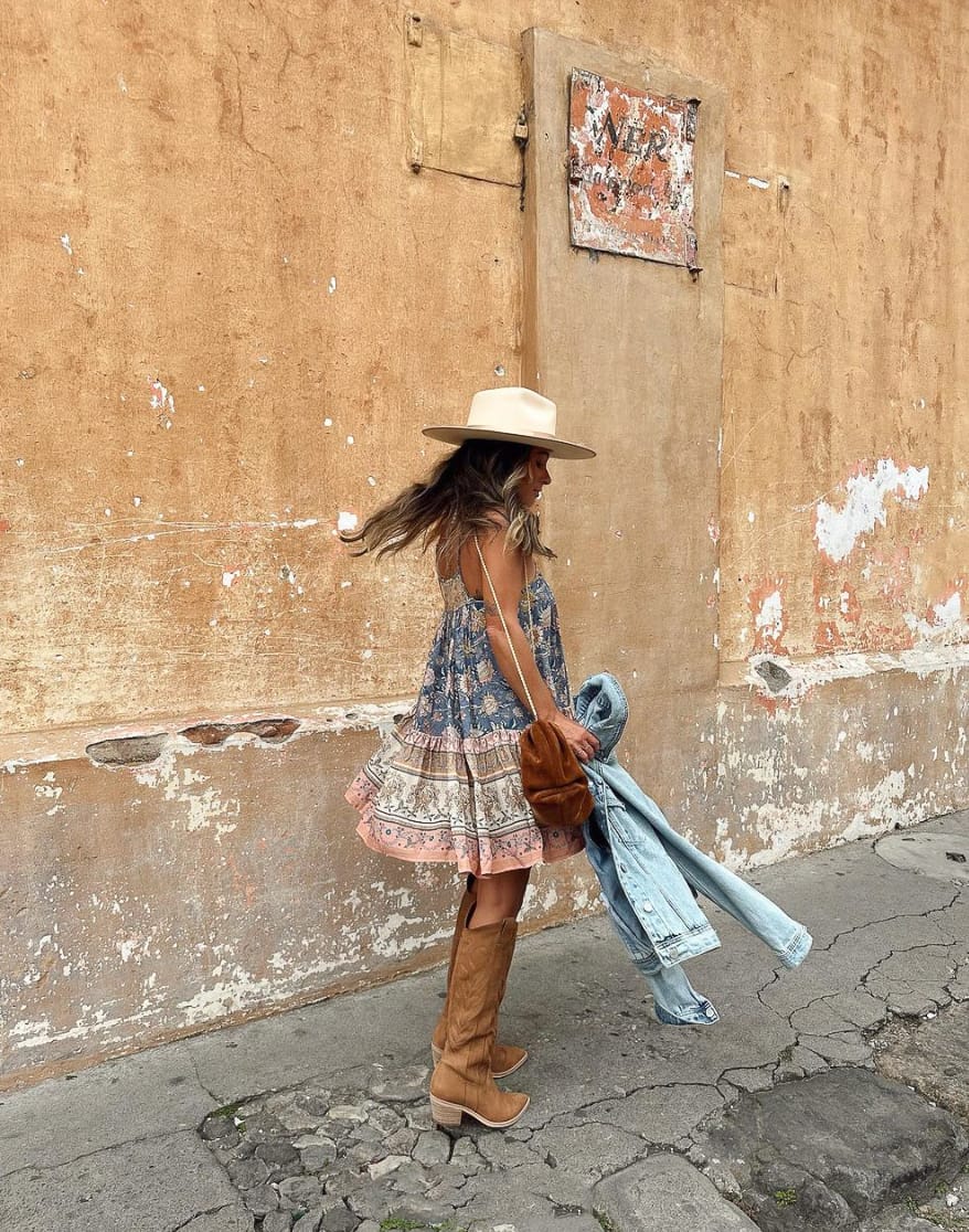 An image of a woman with a boho mini dress, brown cowboy boots, and a white cowboy hat holding a denim jacket
