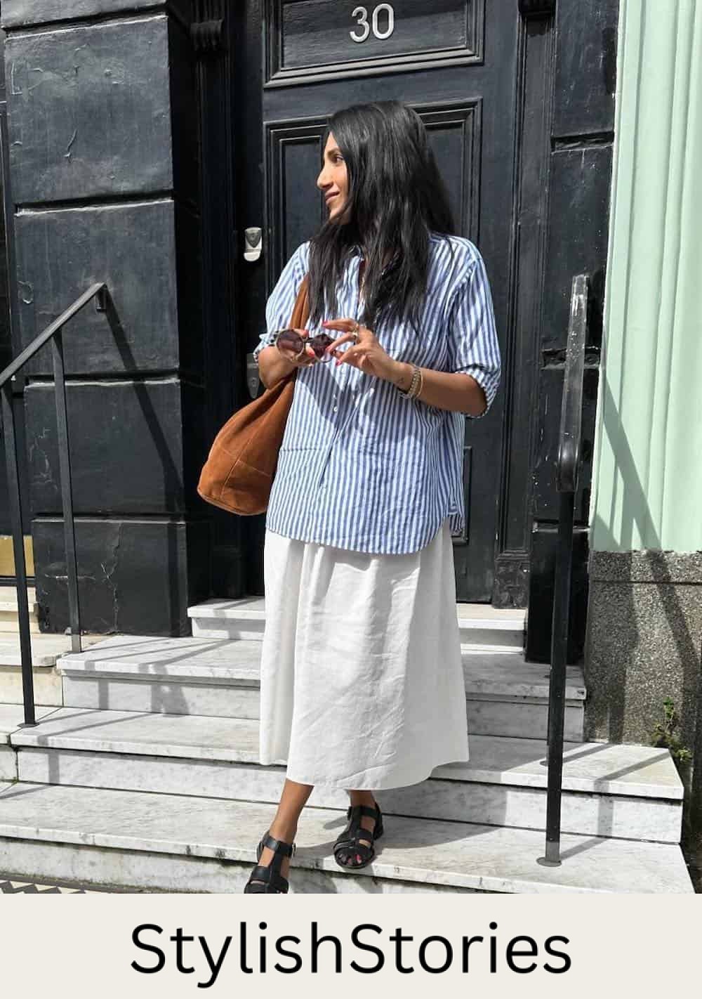 Woman wearing a linen skirt with an oversized blue and white striped button down and sandals.