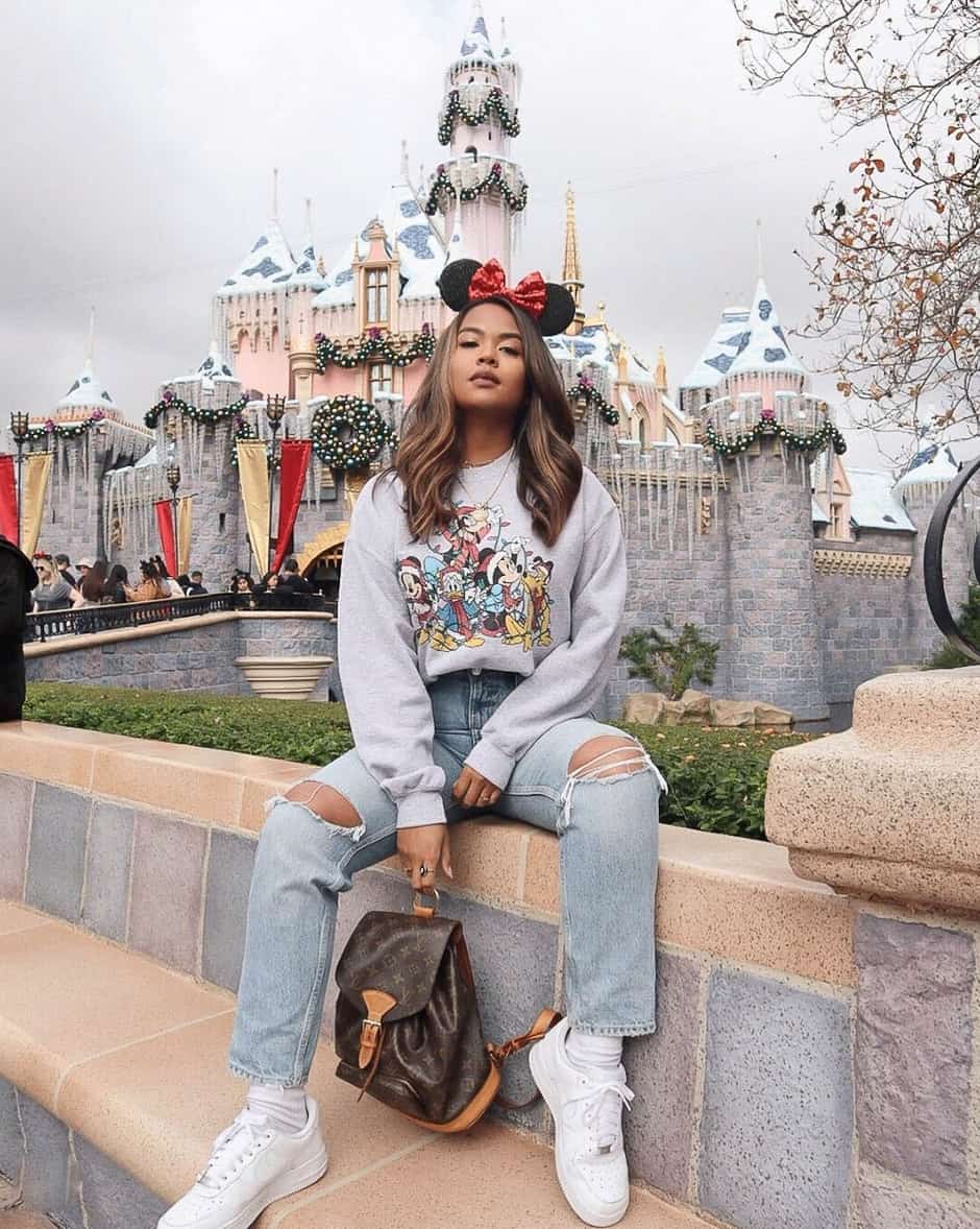 woman at disneyland wearing a graphic sweat shirt, jeans, white sneakers, and black Minnie Mouse ears