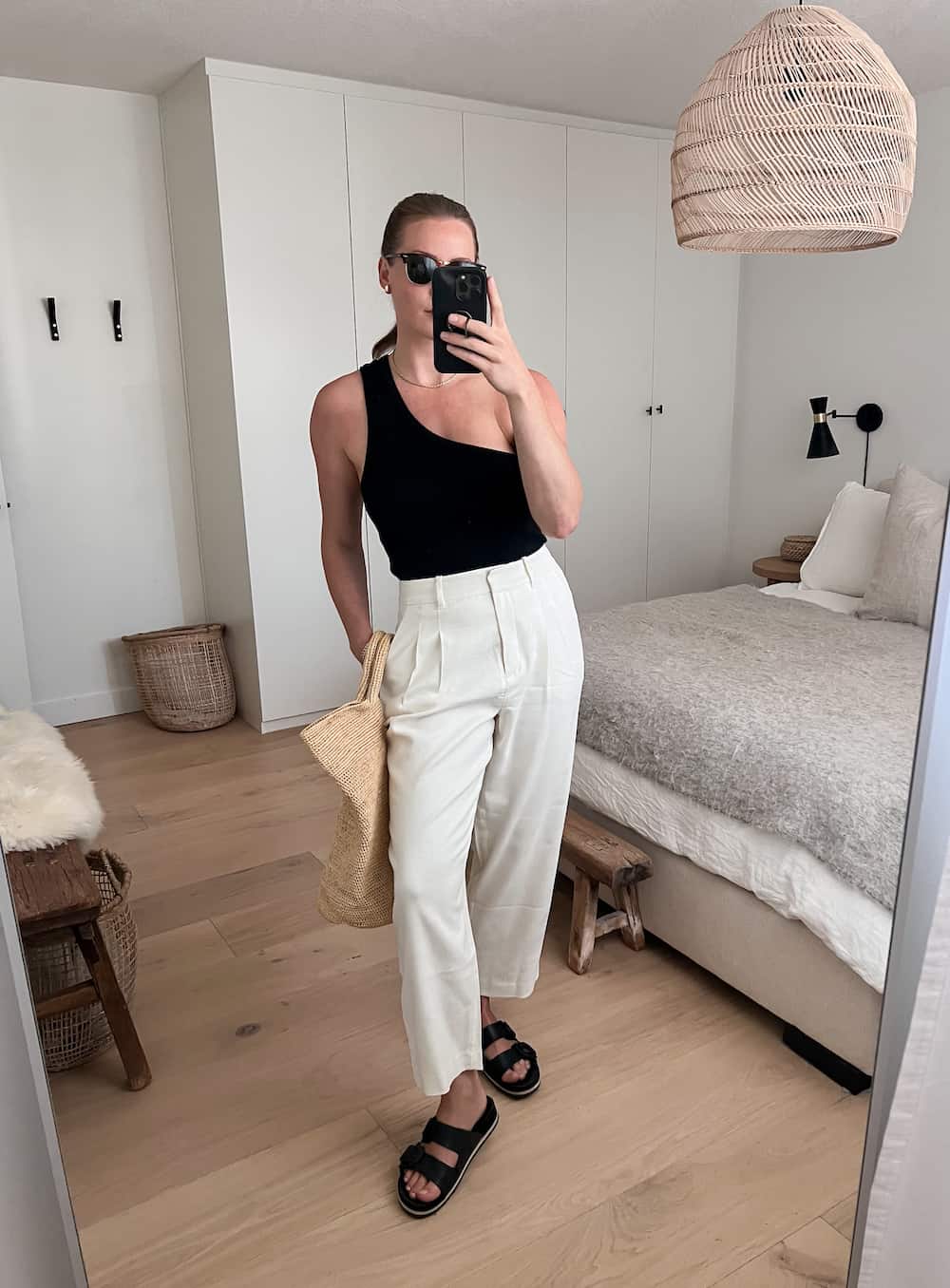 woman wearing a black one shoulder tank, pleated ivory pants, black sandals, and a straw bag