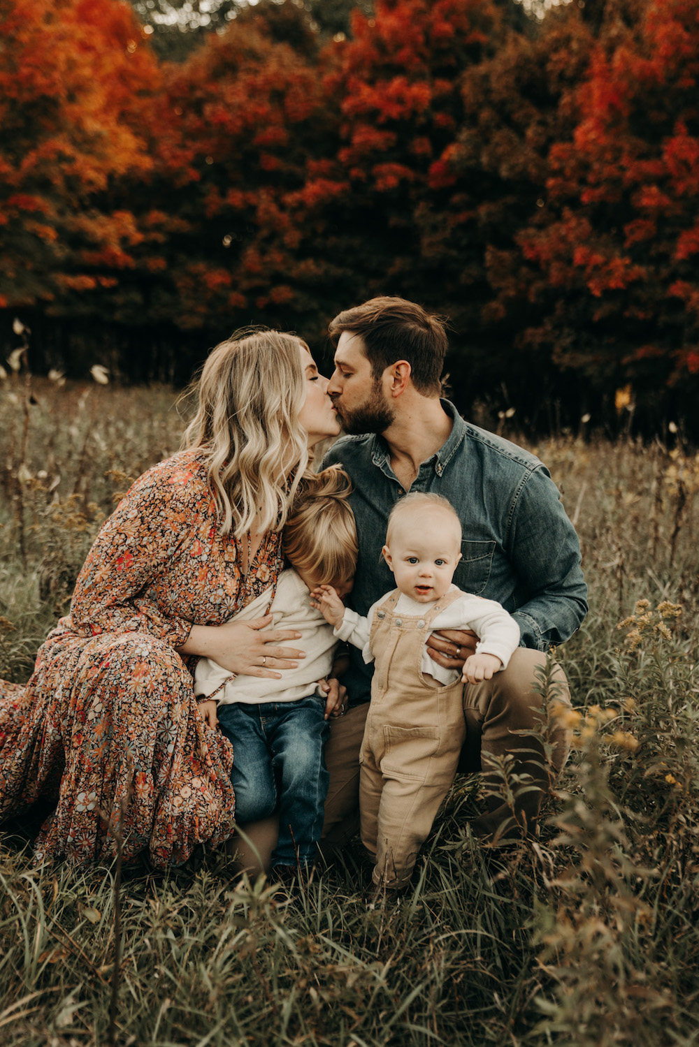 image of a family with a mom, dad, and two young boys in a field in the fall, the mom in a floral dress, and the dad in a denim shirt, the boys in tan and ivory