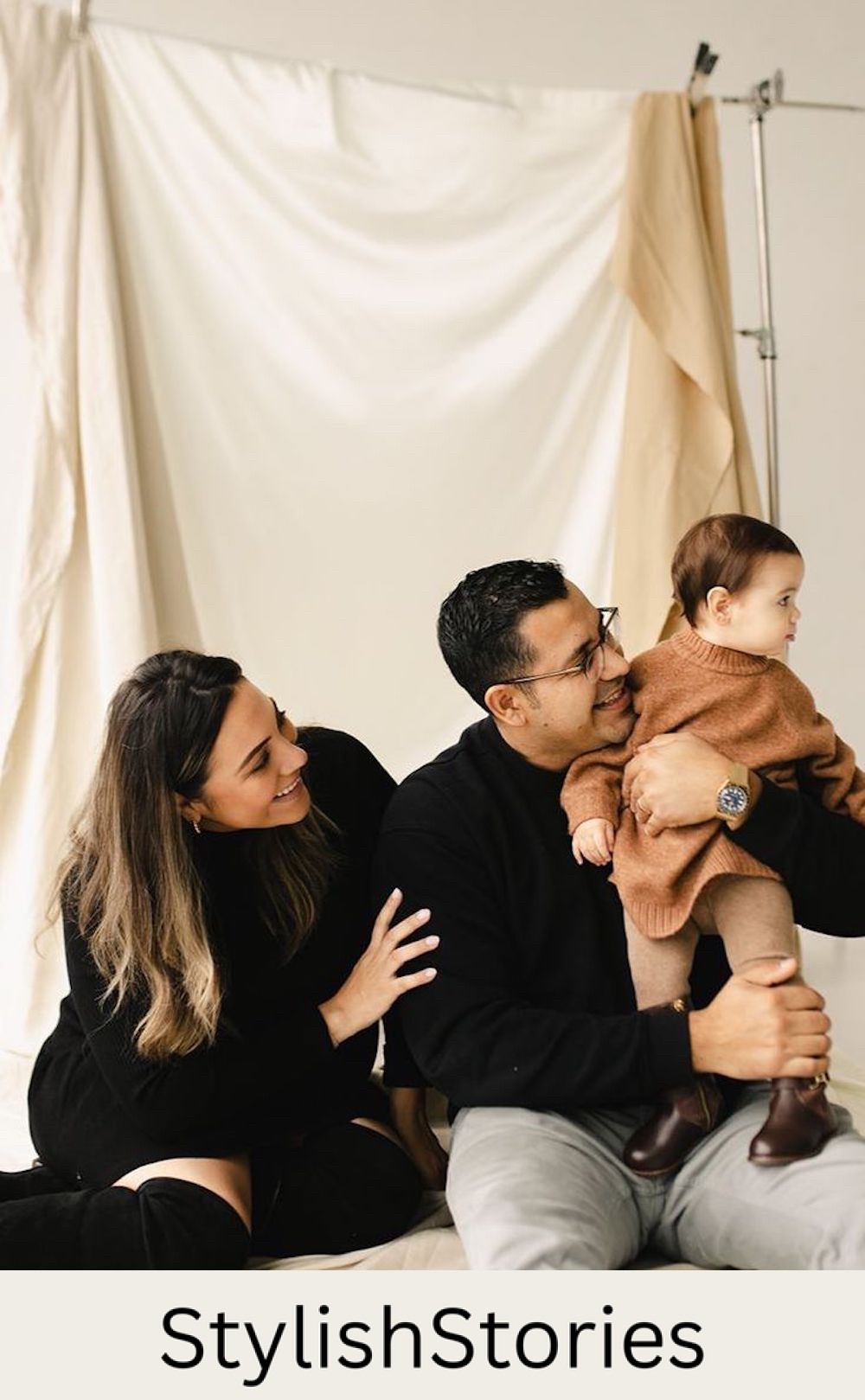 image of a family in a studio in front of a silk backdrop in black outfits with a toddler wearing a brown shirt and pants