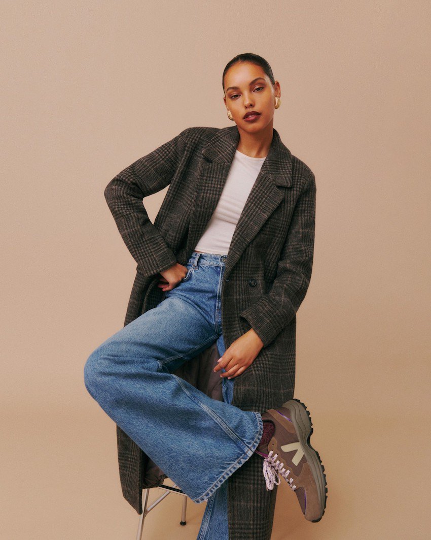 Woman wearing denim wide leg jeans with brown Veja sneakers and a long plaid coat.