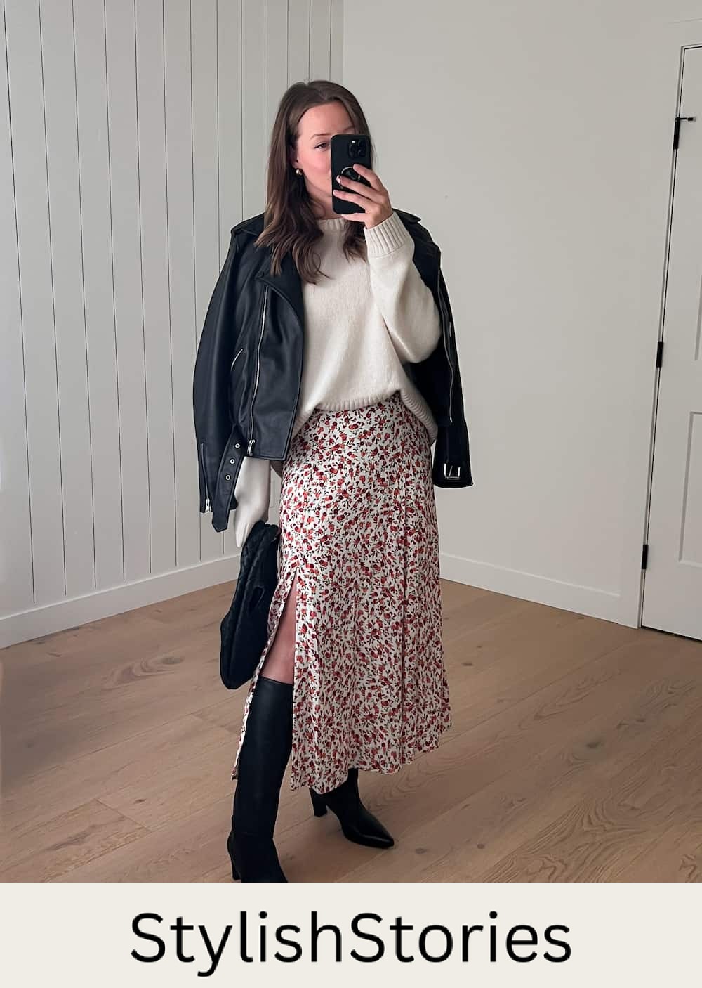 Christal wearing a floral printed dress with a white sweater over and black tall boots. She is also wearing a leather jacket.